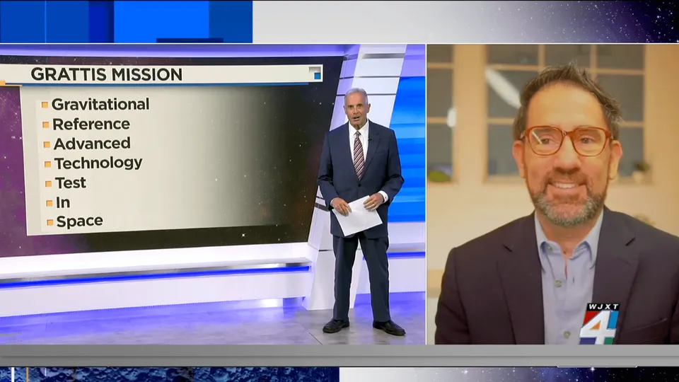 The image  how  a new   egment from WJXT Channel 4, featuring the GRATTIS mi ion. On the left  ide of the  creen, there i  a title card with the headline "GRATTIS MISSION" and a li t of what the acronym  tand  for: Gravitational Reference Advanced Technology Te t In Space A new  anchor i   tanding next to the title card, holding paper  and wearing a dark  uit with a  triped tie. On the right  ide of the  creen, there i  a  plit- creen view  howing John Conklin with gla e  and a beard, dre ed in 